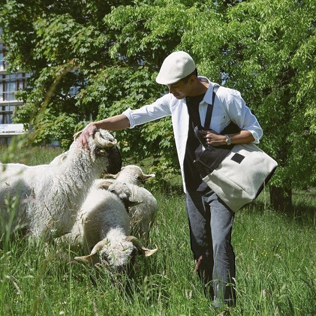 Man wearing a Baabuk BBB Bag in a field with Valais Blacknose sheep, featuring a two-tone wool tote with leather.