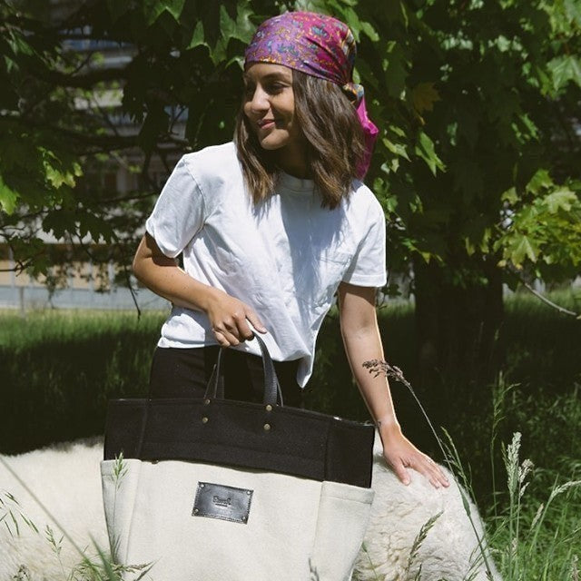 Woman holding a Baabuk BBB Bag in black and cream burel wool with leather handles, standing beside a sheep.