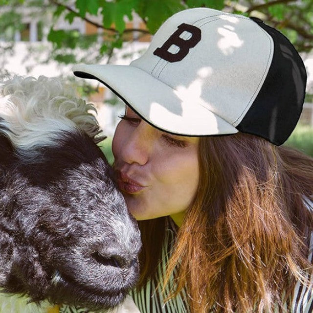 Baabuk B Cap in cream and black wool with embroidered B logo, worn by a woman kissing a Valais Blacknose sheep.