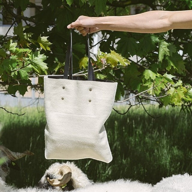 Baabuk Blacknose reversible wool tote bag with leather handles held outdoors near a Valais Blacknose sheep.