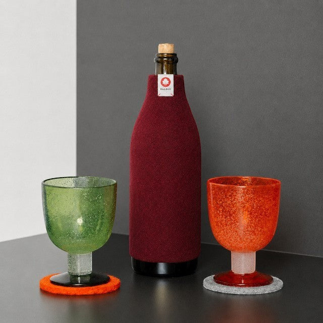 Baabuk bordeaux wool bottle cover insulating a wine bottle next to green and orange textured glasses on a table