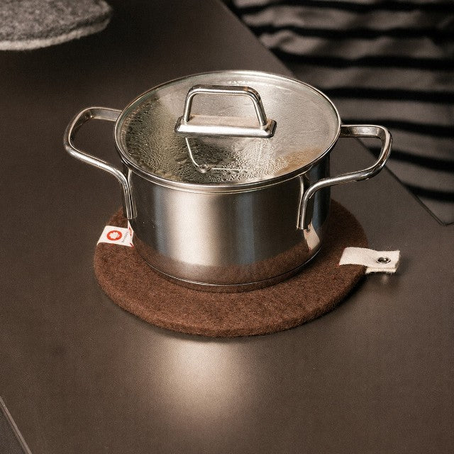 Baabuk brown felted wool trivet hot plate with a leather strap under a stainless steel pot on a kitchen counter.