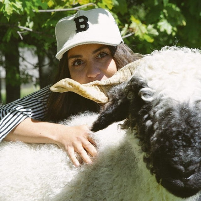 Woman wearing a cream Baabuk wool baseball cap with a black B embroidery next to a Valais Blacknose sheep.