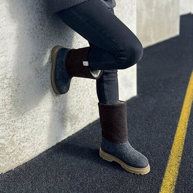 Baabuk Dani dark grey wool boots with brown cuff and rubber sole worn by a person in black jeans leaning on a wall.