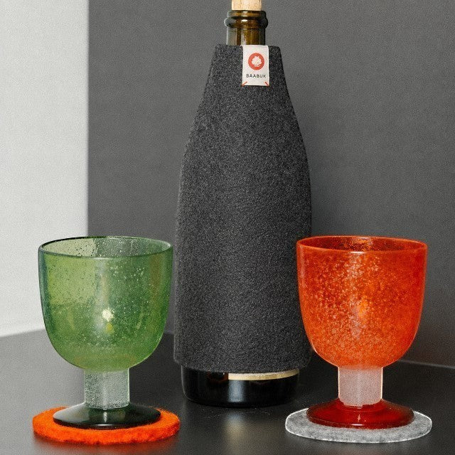 Baabuk dark grey wool bottle cover insulating a glass bottle next to green and orange textured glasses on coasters.