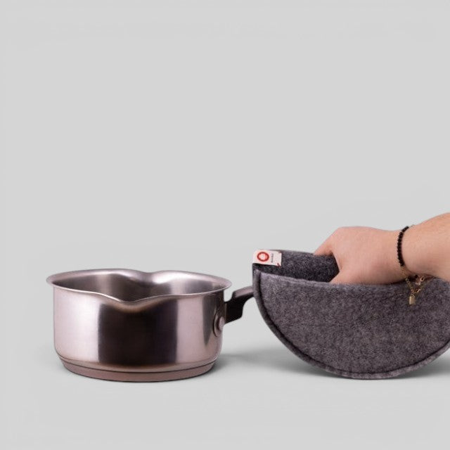 Baabuk dark grey felted wool potholder with a leather strap, held by a hand next to a stainless steel saucepan.