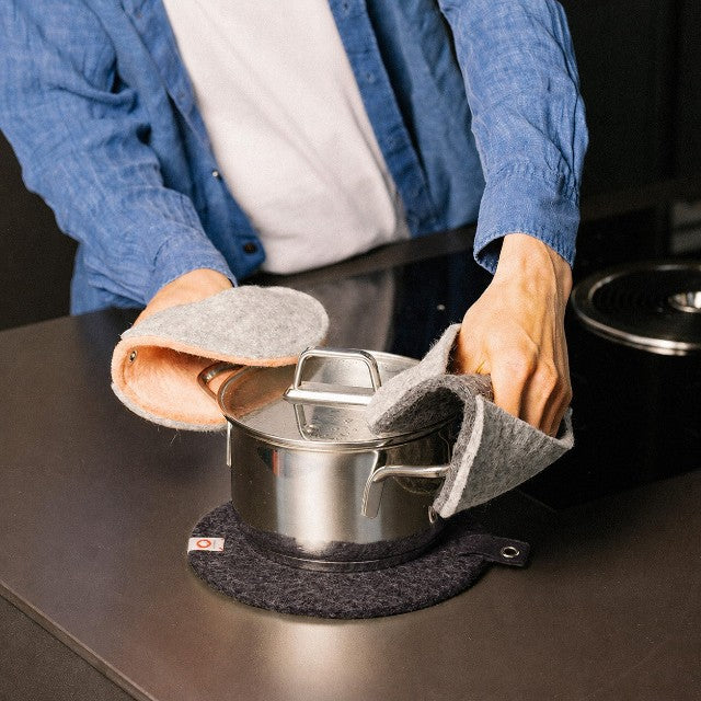 Baabuk dark grey felted wool trivet under a stainless steel pot on a kitchen counter with matching wool oven mitts