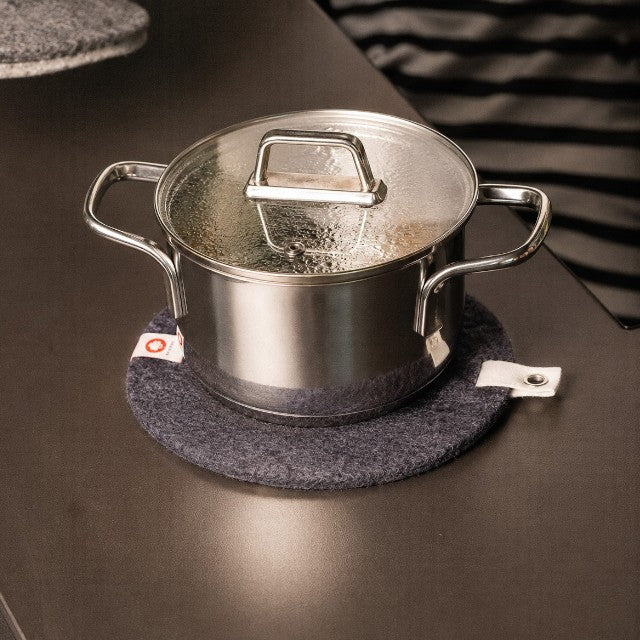Baabuk dark grey felted wool trivet hot plate with a leather strap under a stainless steel pot on a dark surface