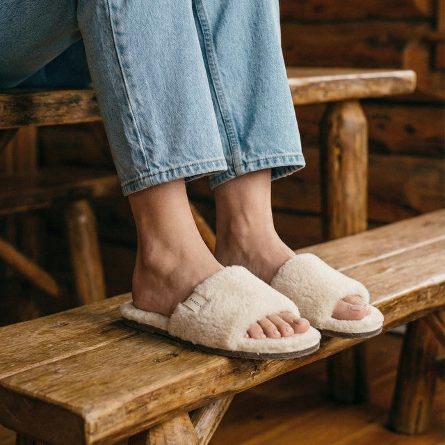 Baabuk Baabulette Fluffy wool slide sandals in cream, worn with blue jeans on a rustic wooden bench.