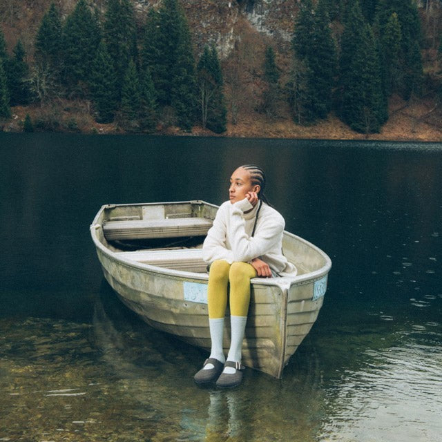 Model wearing grey Baabuk wool Mary Jane sandals while sitting in a rowboat on a mountain lake.