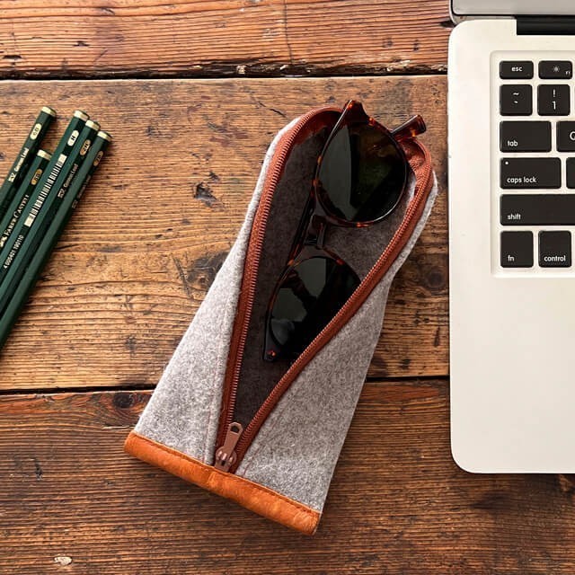 Baabuk grey wool pencil case with brown leather trim and zipper, holding sunglasses on a wooden desk.