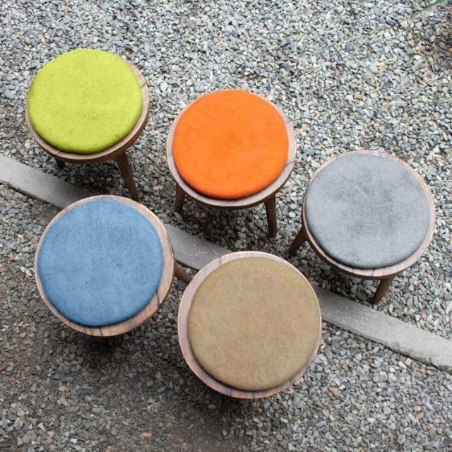 Baabuk handmade wool sitting mats in vibrant green, orange, blue, grey, and brown on wooden stools outdoors.