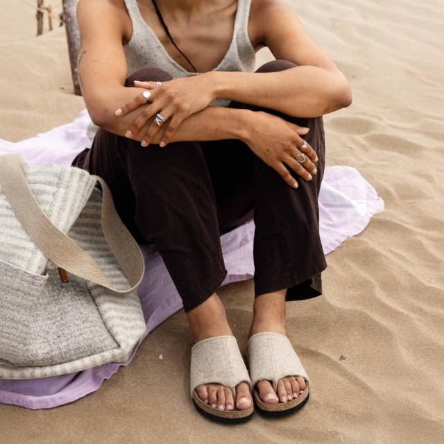 Baabuk Izard Sandal in beige wool with a cork footbed, worn by a person sitting on a sandy beach.