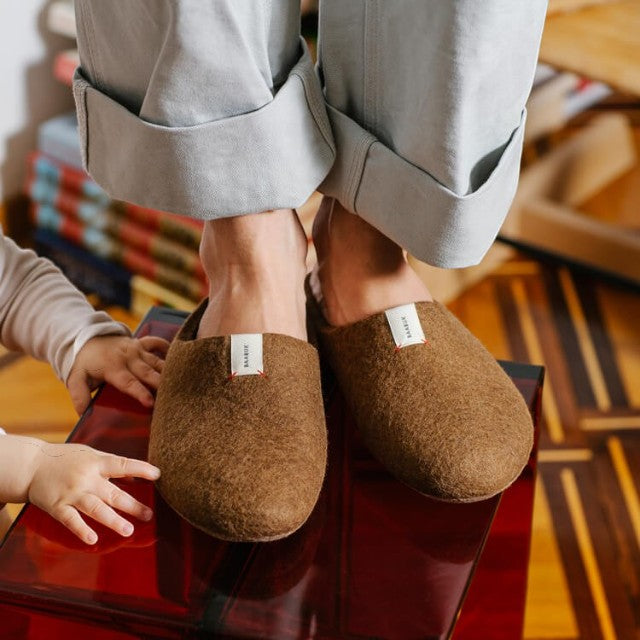 Baabuk Kanga brown wool slippers worn by a person standing on a red stool with a child's hand visible nearby.