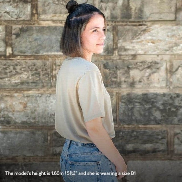 A woman wearing a khaki Baabuk 10-year anniversary merino wool T-shirt, standing against a stone wall.