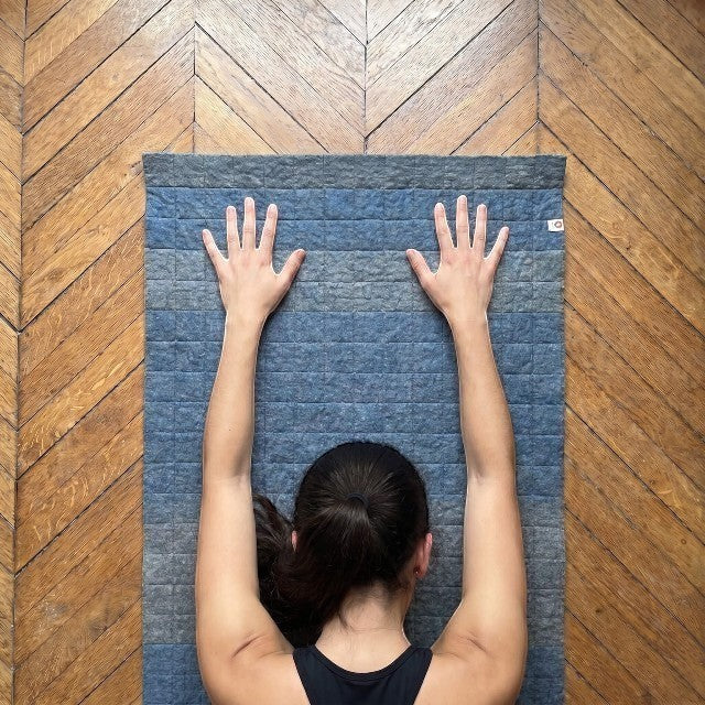 Baabuk light blue felted wool yoga mat with cotton stitching, shown from above with a person in a yoga pose.