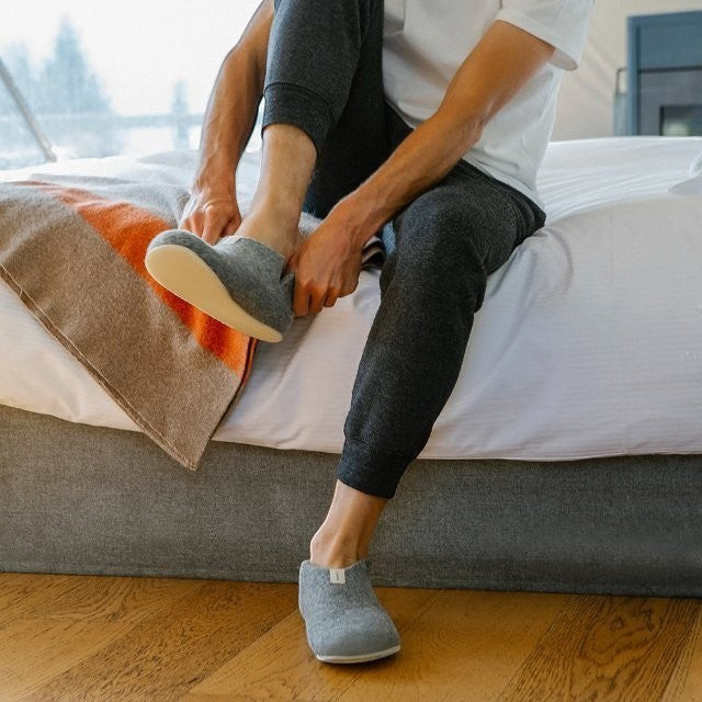 Baabuk Lisa light grey wool slippers with crepe rubber soles being put on by a person sitting on a bed.