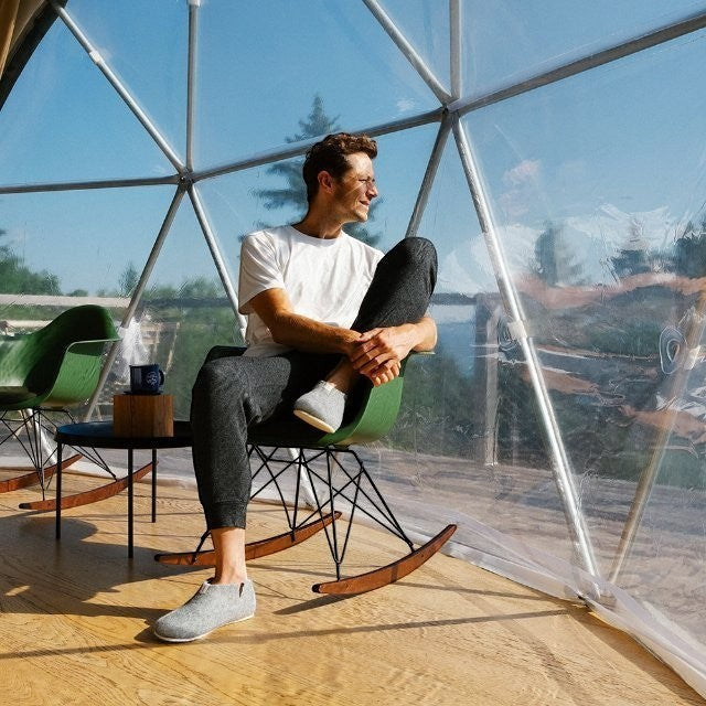 Baabuk Lisa light grey wool slippers worn by a man sitting in a modern rocking chair inside a geodesic dome