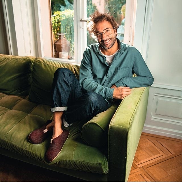 Man wearing Baabuk Mel bordeaux wool slippers while sitting on a green velvet sofa in a sunlit room.