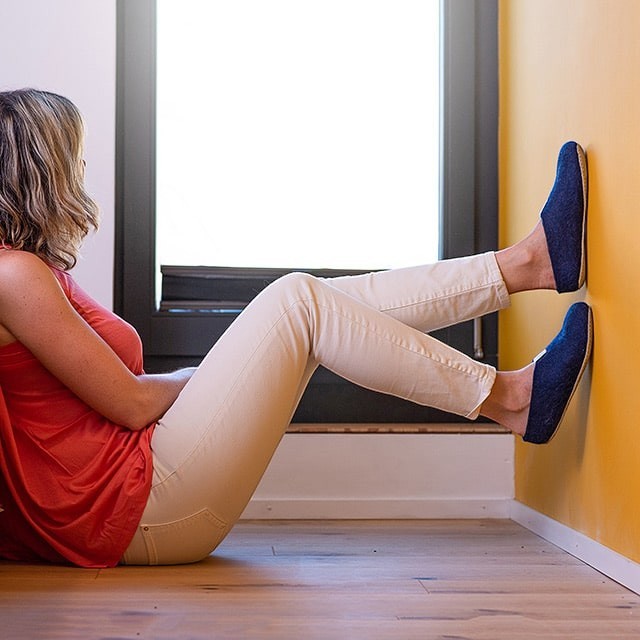 Woman wearing navy blue Baabuk wool slippers with natural rubber soles while sitting against a yellow wall