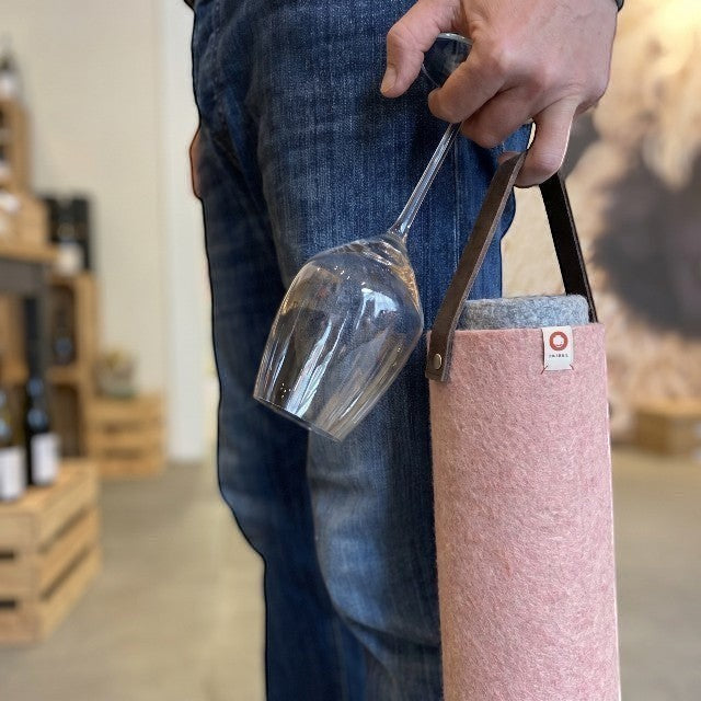Person holding a pink Baabuk wool wine cooler with a leather strap and an empty wine glass in a shop setting.