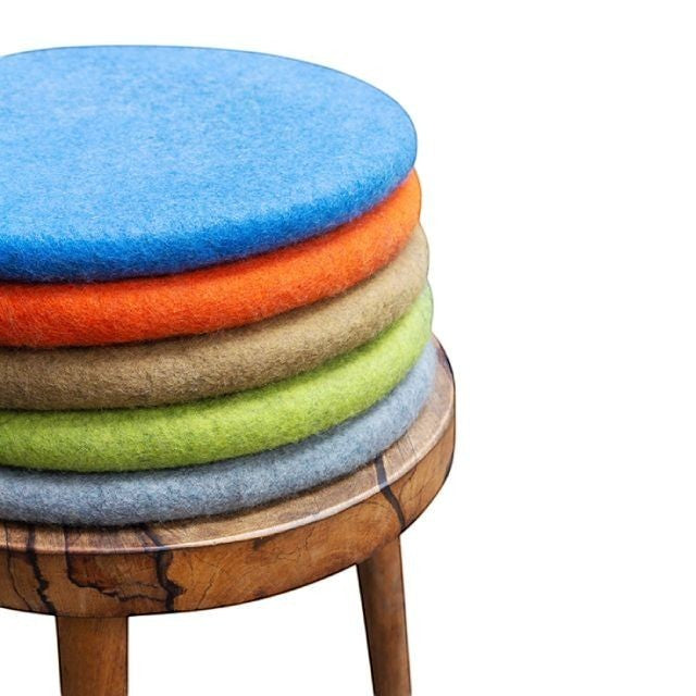 Baabuk handmade round wool sitting mats in blue, orange, beige, green, and grey stacked on a wooden stool.