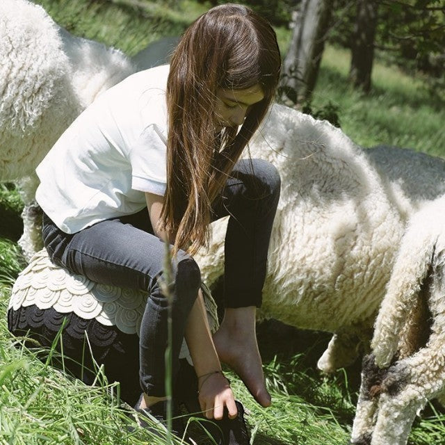 A girl sitting on a Baabuk sheep-shaped stool made of wool in a grassy field surrounded by Valais Blacknose sheep.