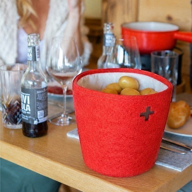 Baabuk Swiss red felted wool basket with a cross cutout, holding potatoes on a dining table.