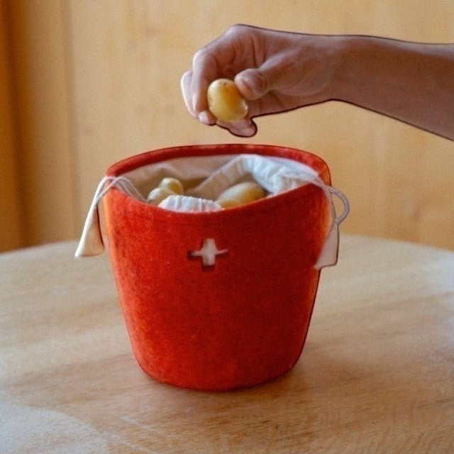Baabuk Swiss red felted wool basket with a cross cutout and drawstring, holding potatoes on a wooden table.