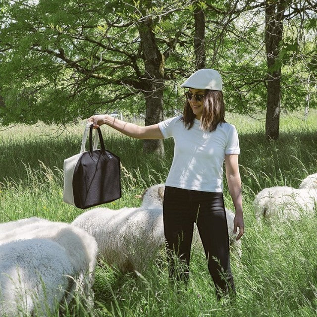 Woman wearing a Baabuk wool beret and holding a two-tone wool bag in a grassy field with sheep.