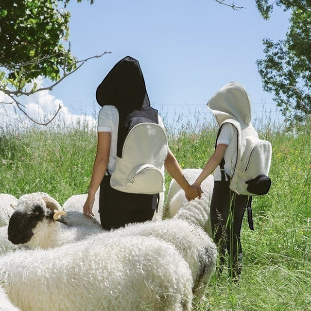 Two people wearing Baabuk wool hooded backpacks while walking through a field with Valais Blacknose sheep.