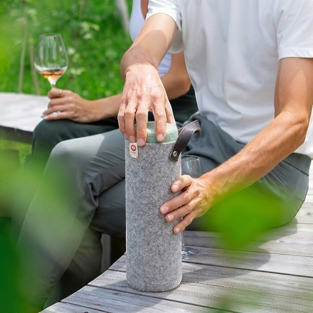 Baabuk light grey wool wine cooler with leather strap, held by a person at an outdoor picnic setting.