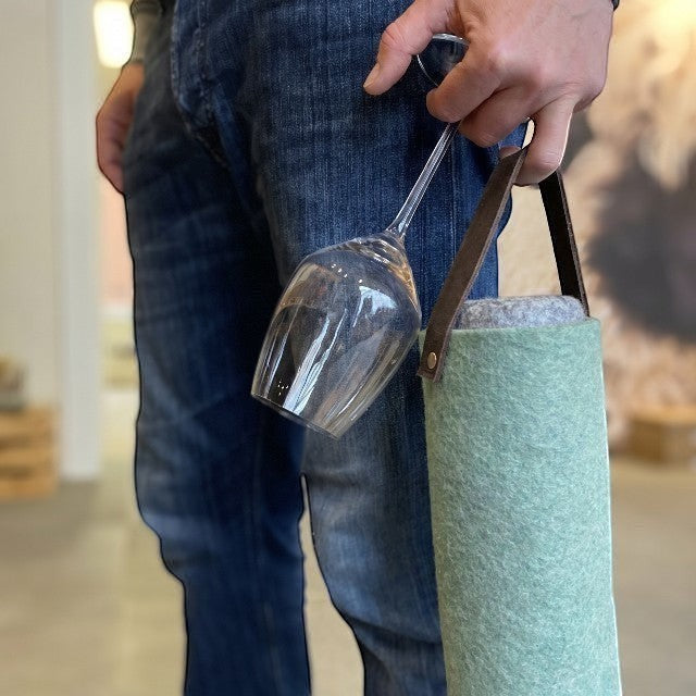 Person holding a turquoise Baabuk wool wine cooler with a leather strap and a wine glass in a casual indoor setting.