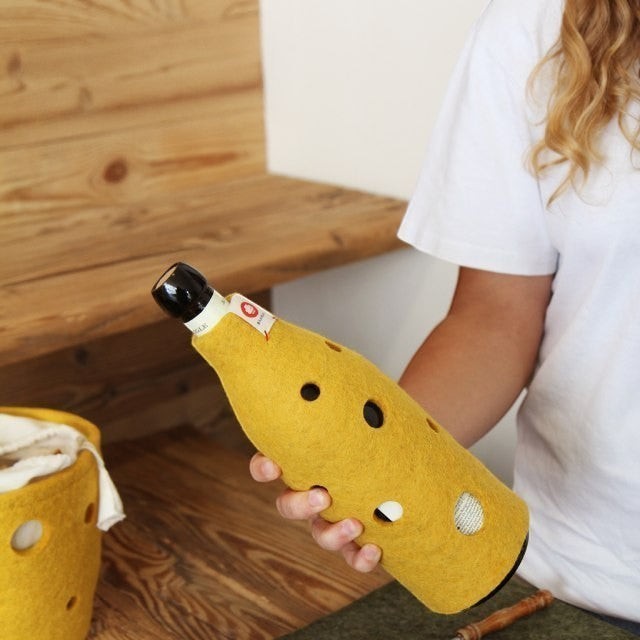 Baabuk yellow wool bottle cover with Swiss cheese hole design held by a person in a wooden interior setting