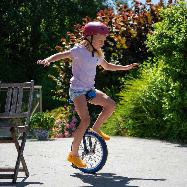 A young girl wearing yellow Baabuk Yves Kids wool slippers while riding a unicycle outdoors in a sunny garden.