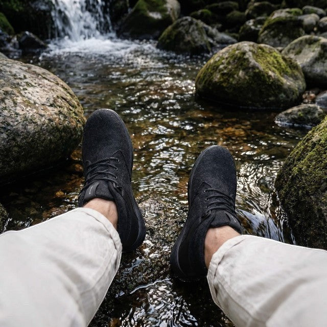 Baabuk Zero Wooler High black wool barefoot shoes worn by a person sitting on rocks by a flowing stream